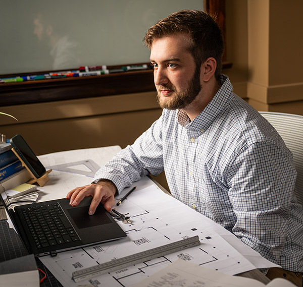 Jonathan Bonamarte sitting at a desk with design drawings while using a laptop.