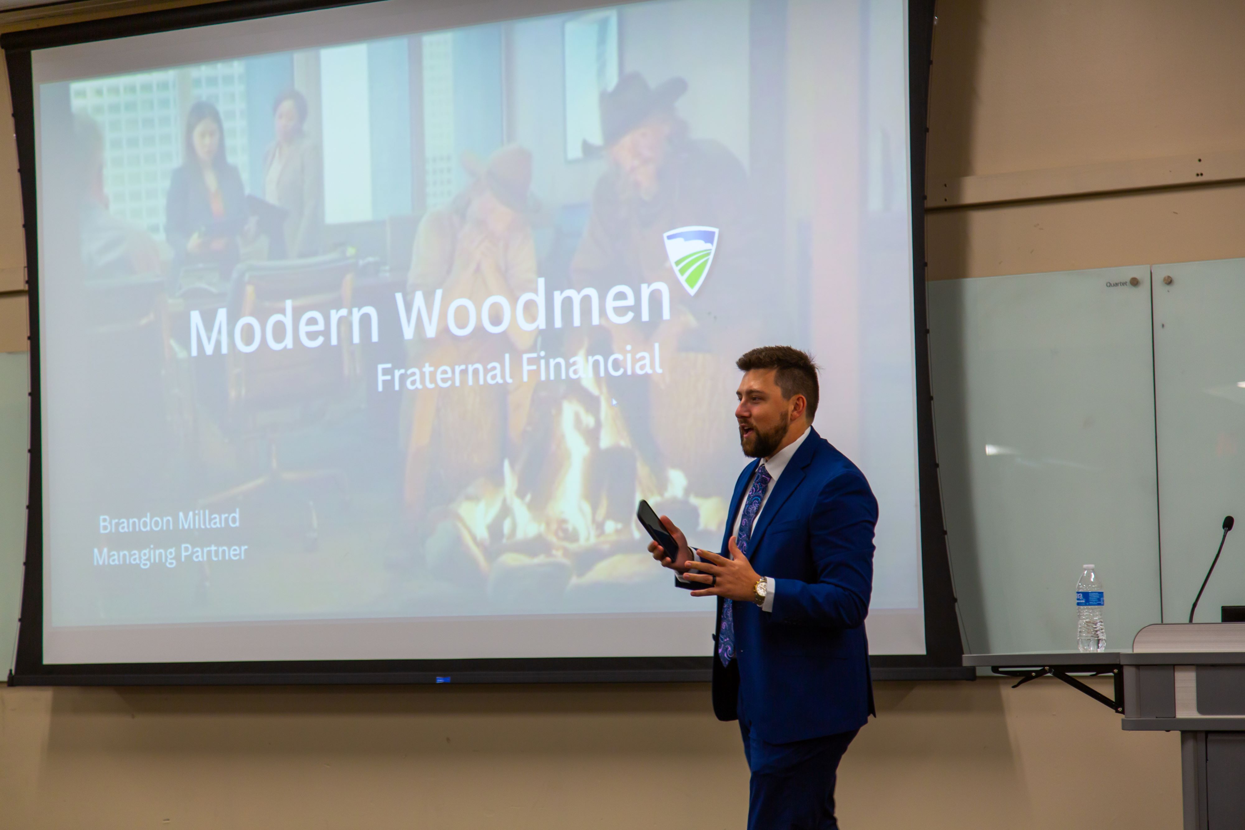 A guest speaker in a blue suit presents in a classroom in front of a projected slide reading “Modern Woodmen Fraternal Financial.”