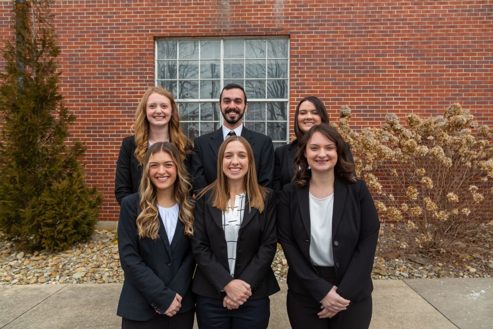 Group of seven College of Business students in professional attire standing outdoors in front of a brick building on campus.