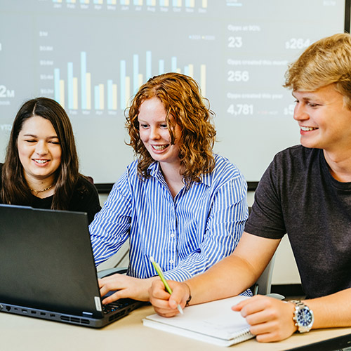 Three students sitting at a desk looking at a laptop with bar graphs being projected in the background.