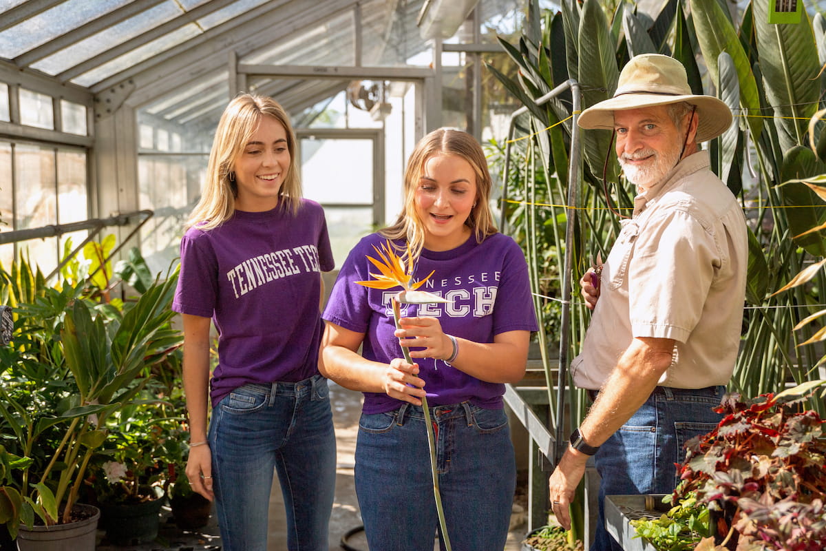 Students examine a tropical plant with an instructor during a hands-on horticulture lesson in a greenhouse.