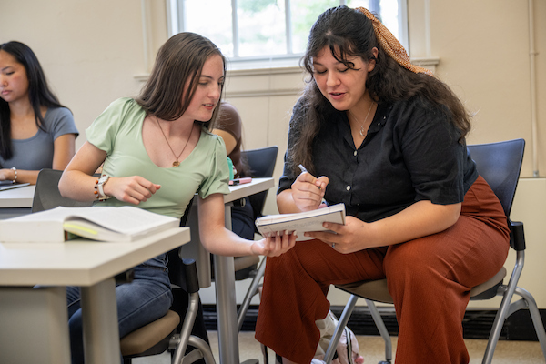 Two students review notes together in a classroom, one writing in a notebook while the other holds it.