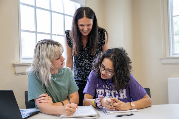 An instructor leans over a classroom table to help two students working with notebooks beside a laptop.