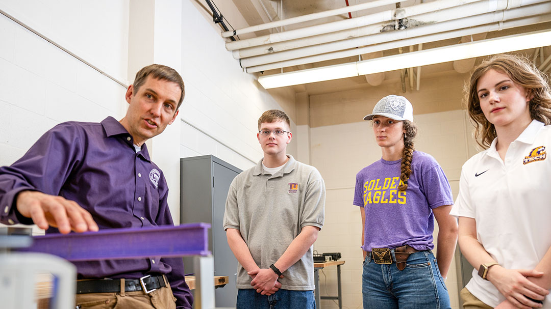 Engineering students observe a faculty member demonstrating structural testing equipment in a laboratory.