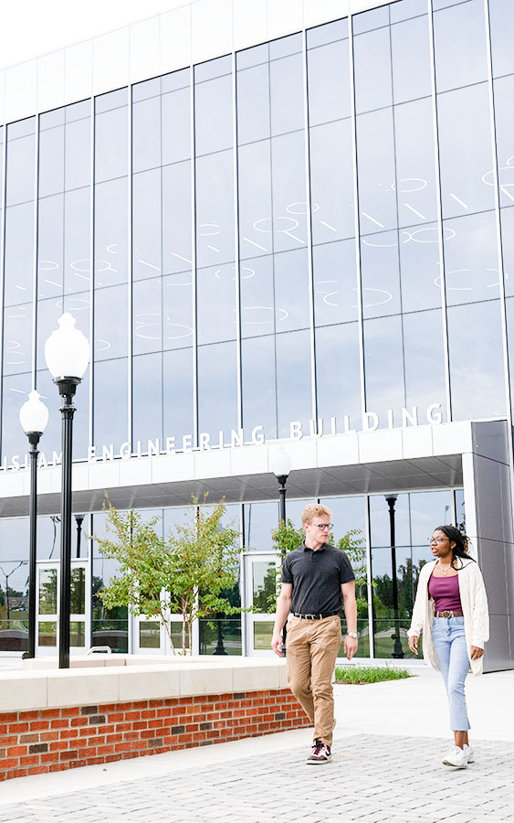 Two students walk in front of the glass-fronted engineering building on campus.