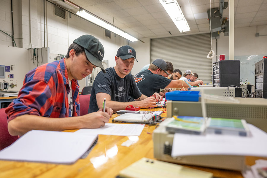 Engineering students work at electronics lab stations, wiring and testing circuits while taking notes during a hands-on laboratory class.