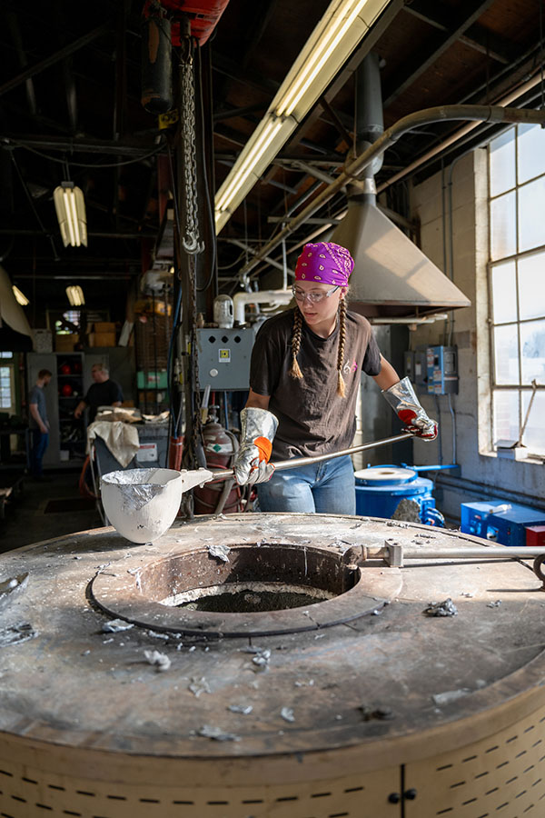 Engineering student wearing safety gear uses a long-handled tool beside an industrial furnace in a manufacturing laboratory.