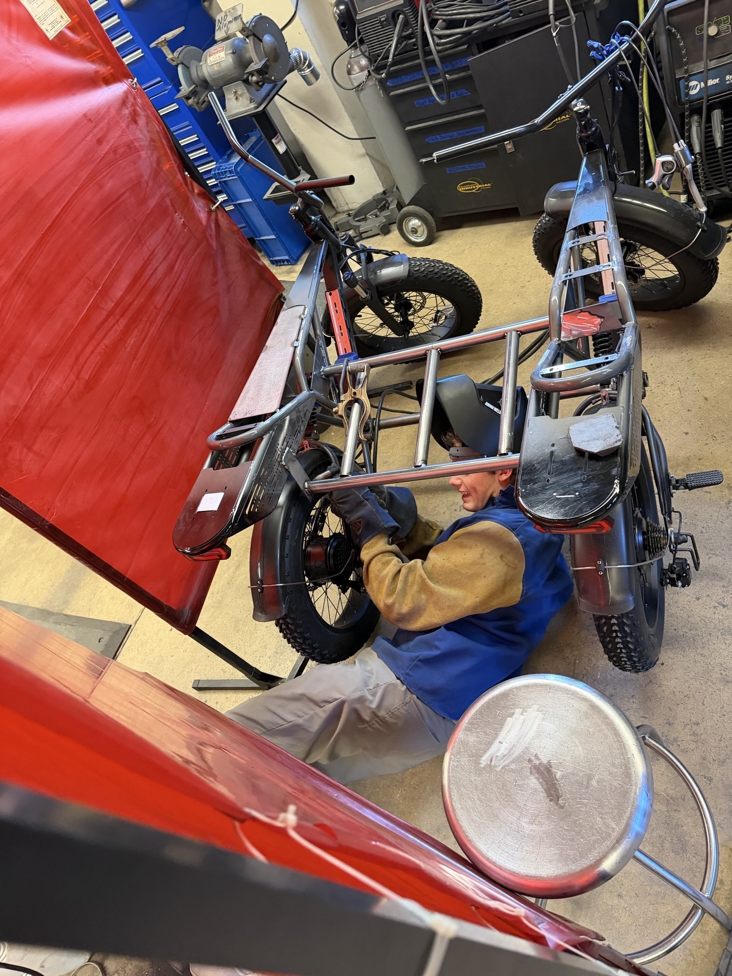 A student is seen on the ground tinkering with a wheelchair mounted above him. 