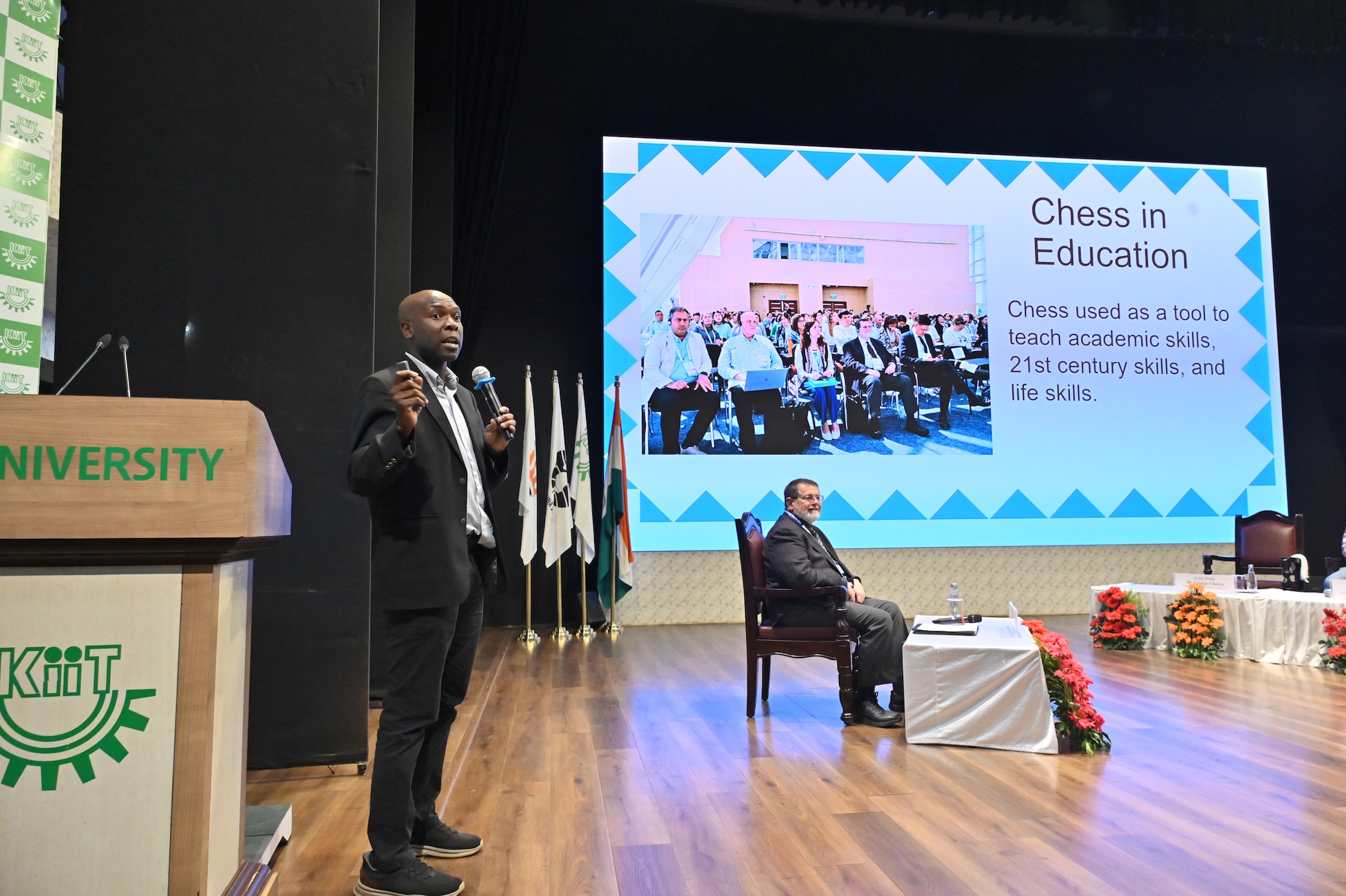 George Chitiyo speaks on stage holding a microphone and wearing a suit. 