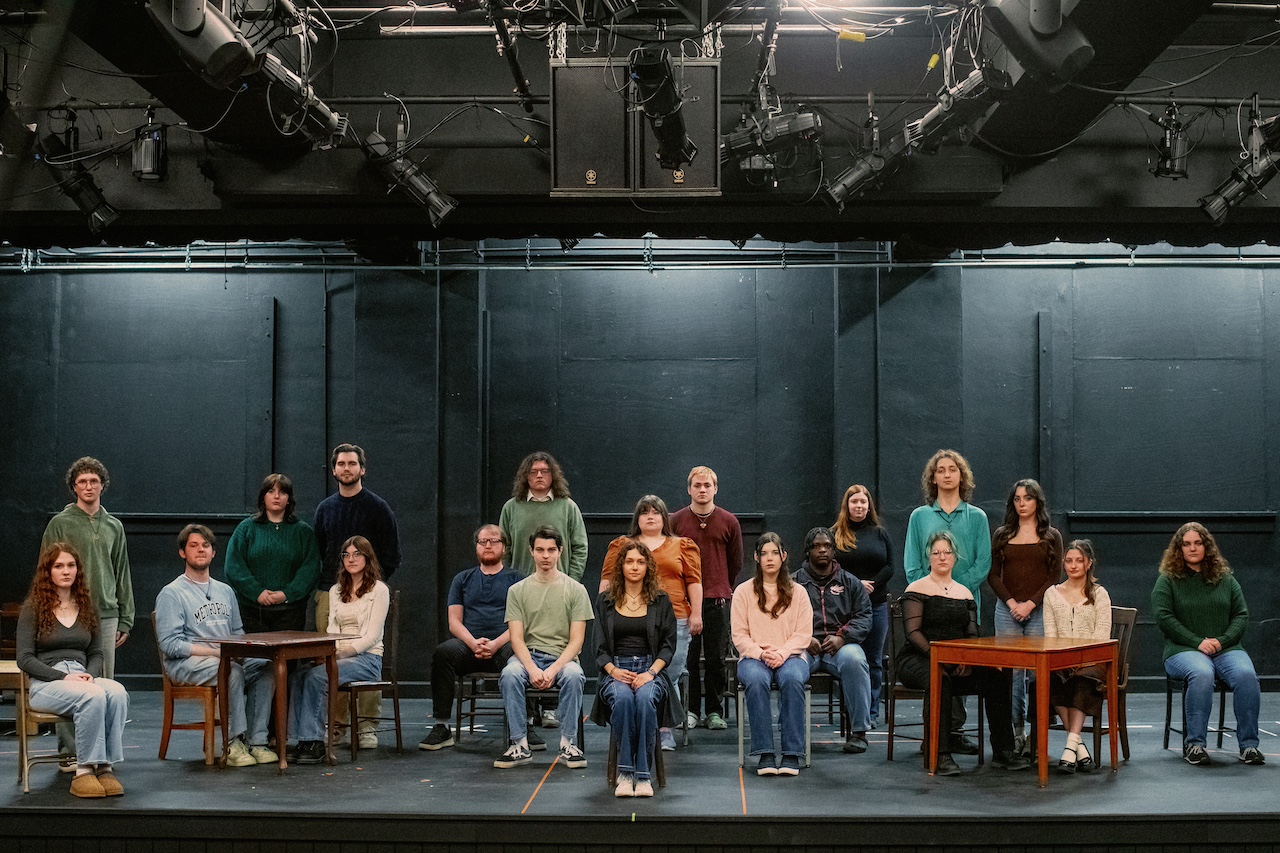Cast members sit and stand looking directly at the camera against a black backdrop. 