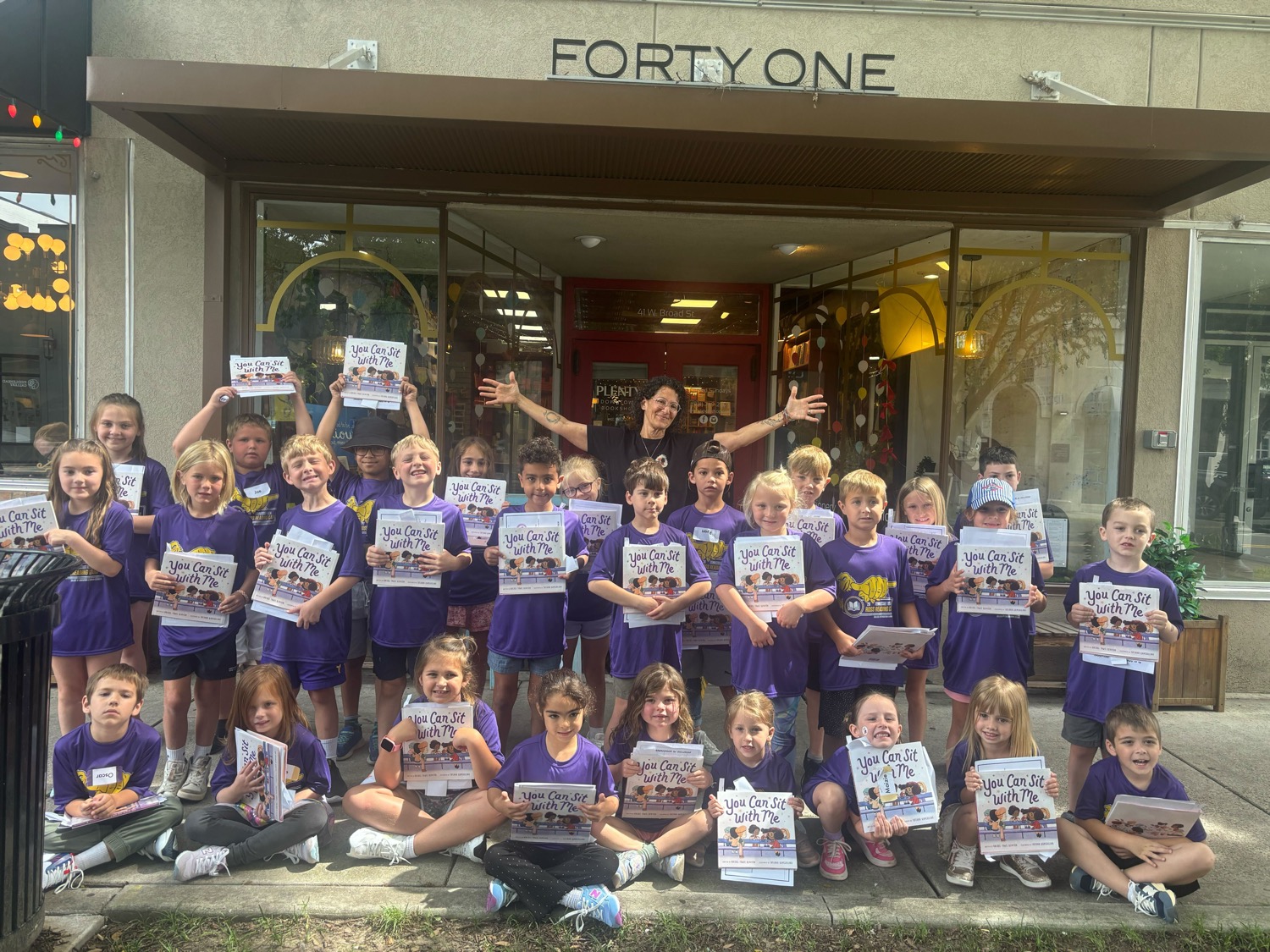 A group photo of students smiling holding up books. The author stands and smiles in the background.