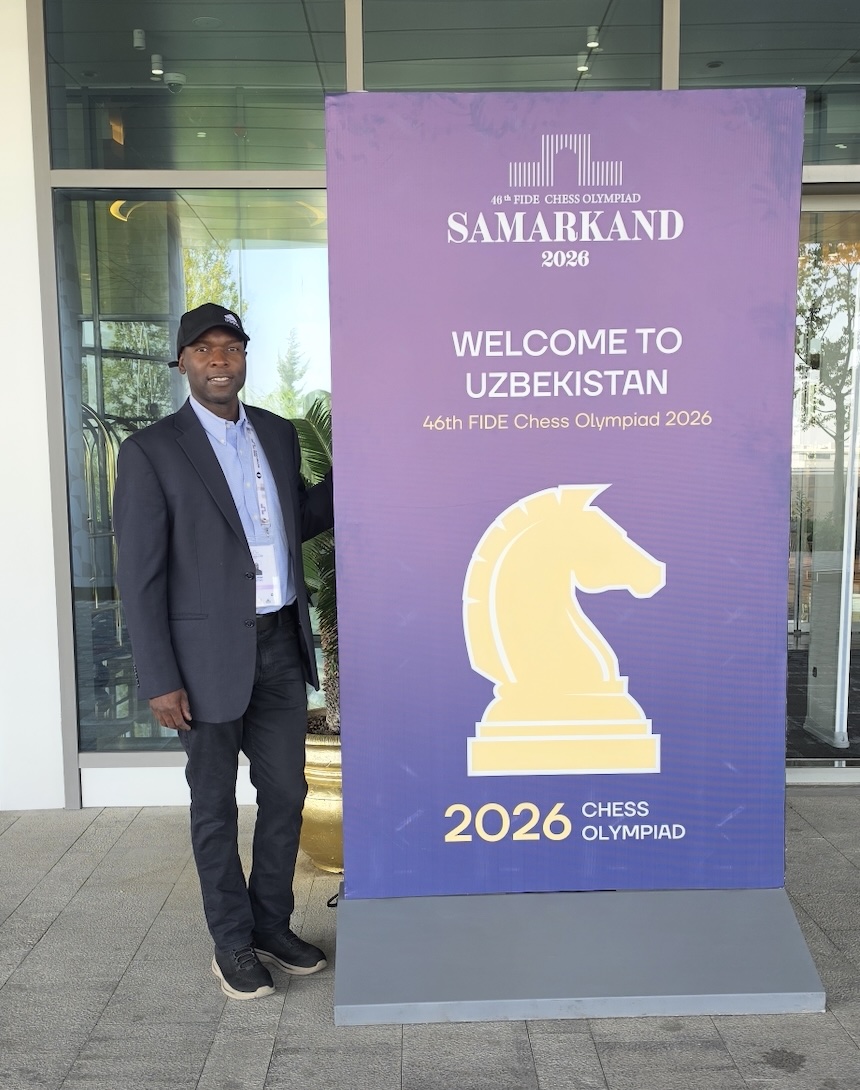 George Chitiyo is pictured in a suit and hat standing next to a large sign for the chess summit
