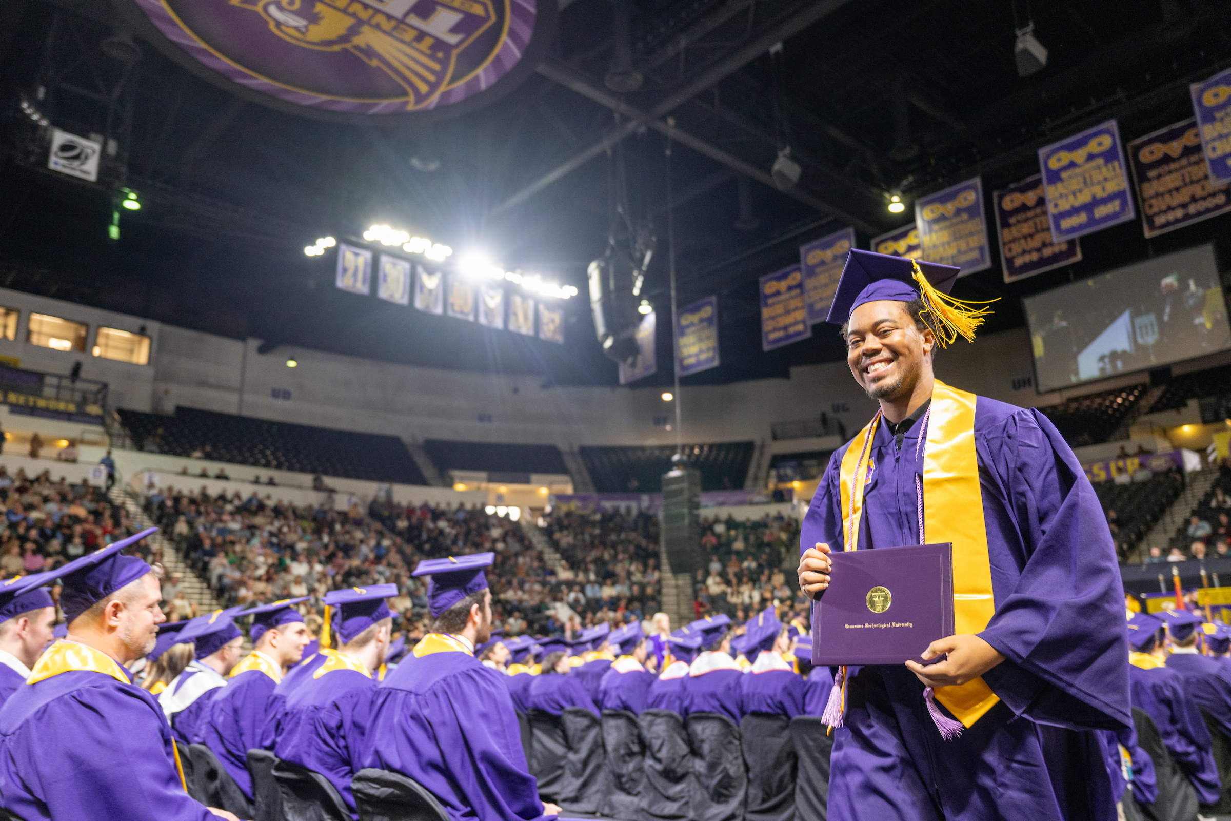 a male-presenting student of color walking at Commencement, smiling and displaying their diploma