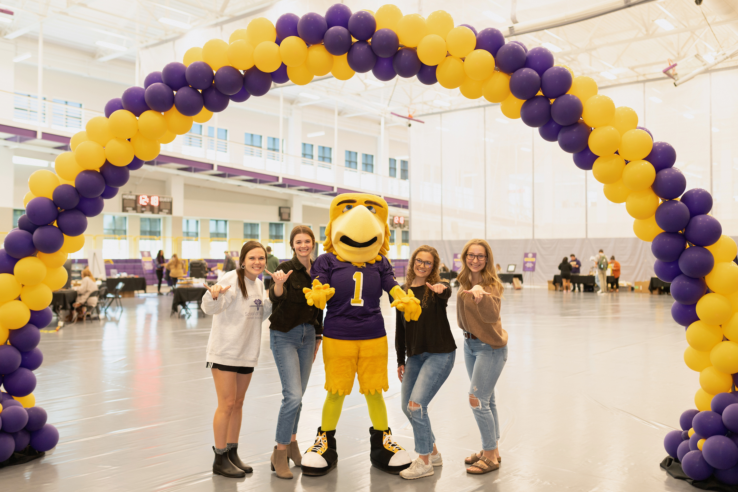 Awesome and four female-presenting students giving the Wings Up hand sign beneath a purple and gold balloon arch