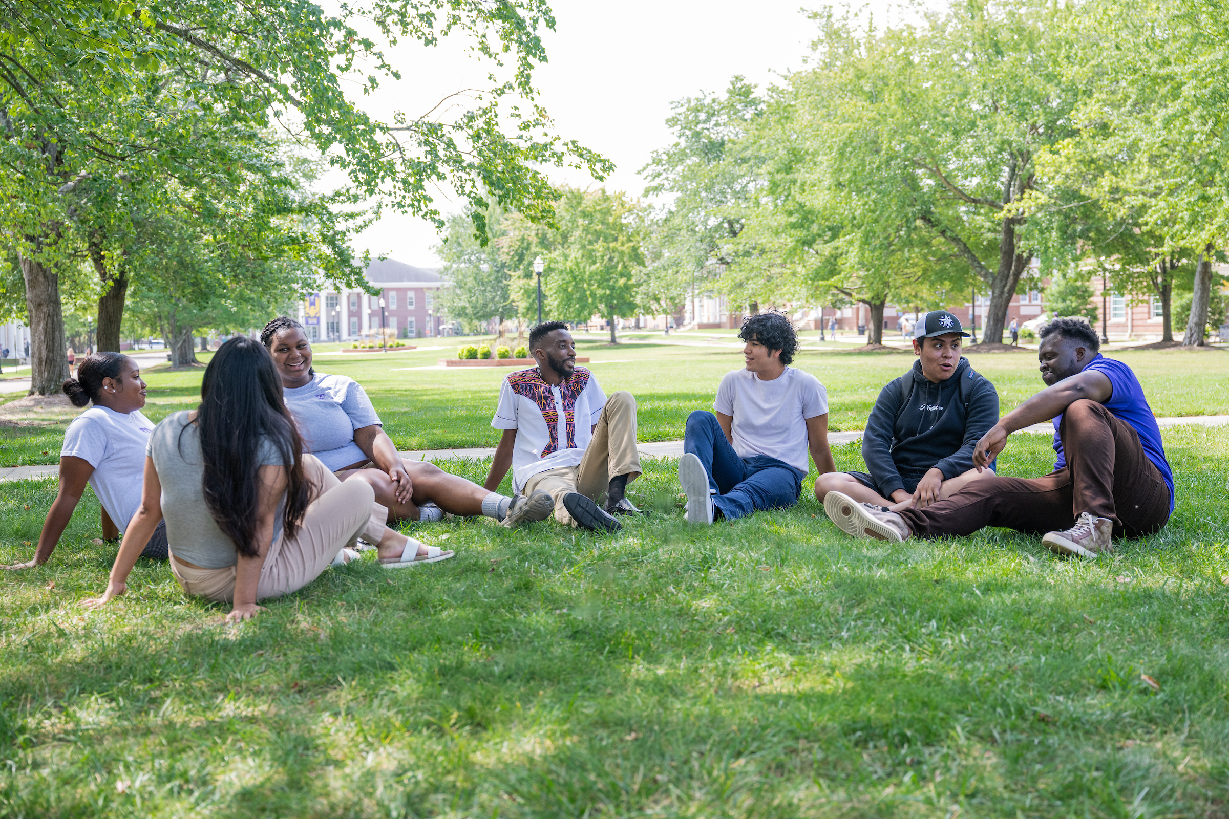 students sitting together on the main quad lawn beneath green shade trees, with Derryberry Hall faintly visible in the background