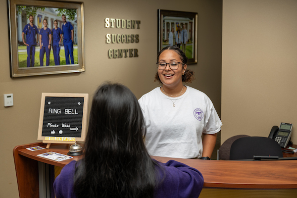 A smiling staff member welcomes a student at the reception desk of the Nursing Student Success Center.