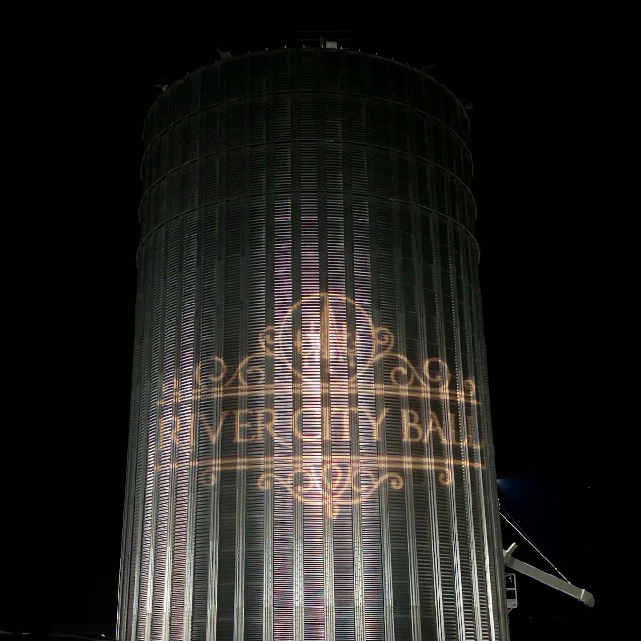 The River City Ball 2026 logo projected onto a towering grain silo at Catesa Farms during the annual community fundraising gala, illuminated against the night sky.