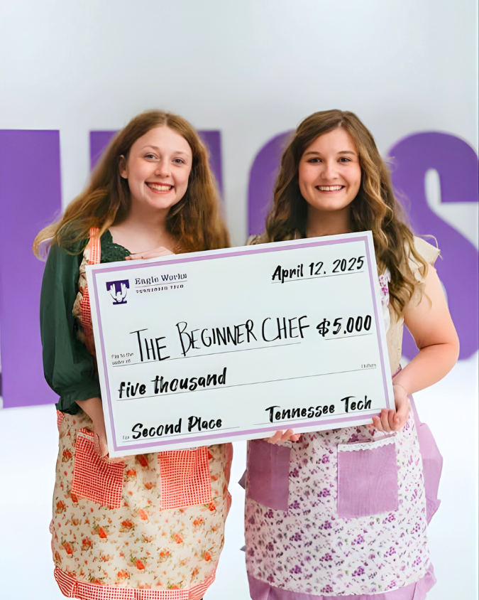 Two Tennessee Tech students stand side by side smiling and holding a large ceremonial check. The check reads “The Beginner Chef – $5,000” and “Second Place – Tennessee Tech,” dated April 12, 2025. They are wearing patterned aprons, representing their student venture, and standing in front of a purple Eagle Works backdrop