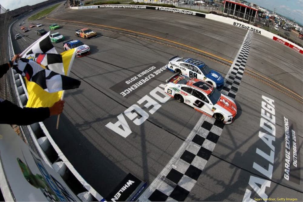 A fisheye view of the finish line at Talladega Superspeedway. The number 9 car crosses the checkered finish line just ahead of car number 88. The checkered flag waves in the left foreground of the photo.