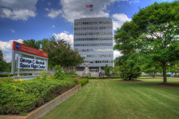 The outside of the George C. Marshall Space Flight Center - there is a mowed green lawn and a tall building with the American flag on top. 
