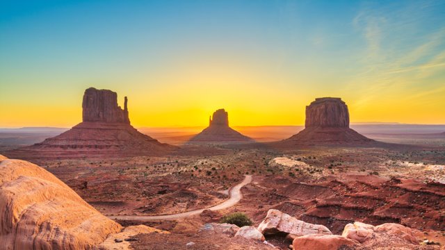 A sunrise over the Utah desert. There are four buttes in the distance and layered rock in the foreground.