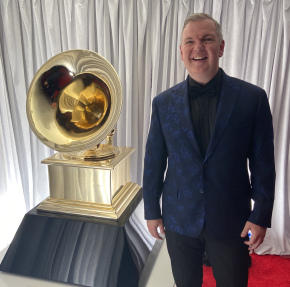Craig Terry, wearing a dark blue damask suit jacket and smiling, stands beside a large replica of a grammy award