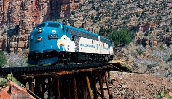 A blue Verde Canyon Railroad train with the number 1512 goes across a bridge with an outcrop of red and brown sandstone behind it.