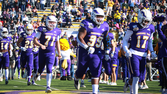 Football players run onto the field in Tucker Stadium. The stands behind them are full of onlookers wearing gold and purple.