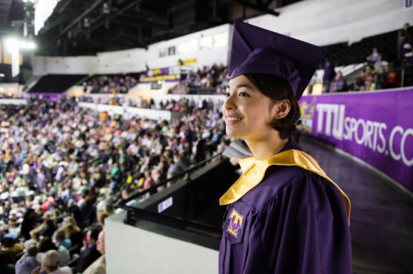 A female student in her purple cap and gown stands at the top of Hooper Eblen smiling at the crowd gathered for commencement.