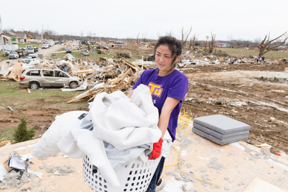 A female student carries a laundry basket of bedding. Threre are piles of wood and debris behind her  - what remains of homes destroyed by the tornadoes.