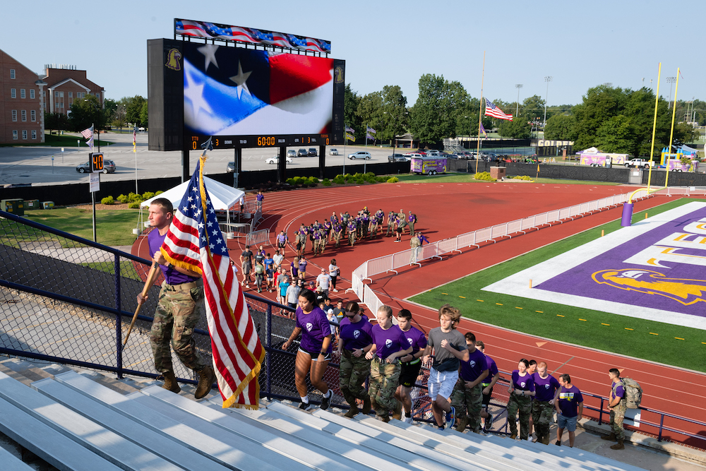 A portrait of ROTC students running up the stairs of Tucker Stadium. The student in front is carrying an American Flag.